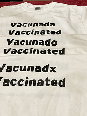 A close up of three folded white tshirts layered vertically on top of each other. The top one reads Vacunada Vaccinated. The middle one reads Vacunado Vaccinated. The bottom one reads Vacunadx Vaccinated.